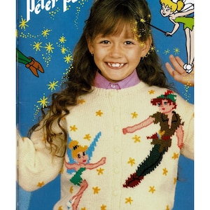 May include: A cream-coloured knit cardigan featuring a button-down front and intarsia designs of Peter Pan and Tinker Bell. The jumper is decorated with small yellow star motifs scattered across the front, set against a blue background with text.