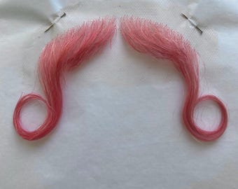 May include: Two pink fake mustaches on a white background. The mustaches are attached to a white mesh material with pins.
