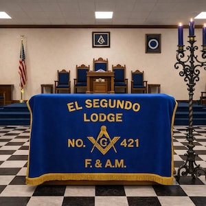 May include: Interior shot of a Masonic lodge room. A blue table cloth with gold fringe displays the text "EL SEGUNDO LODGE" and other Masonic symbols. An American flag stands in the background, along with chairs and a candelabra.