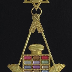 Puede incluir: Colgante masónico dorado con un diseño de estrella, compás y escuadra. El colgante presenta una estrella central con un emblema, un juego de compases y una escuadra, con gemas rectangulares de colores. El fondo es negro.