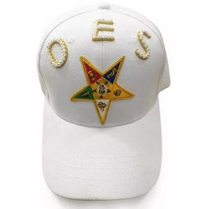 Puede incluir: Gorra de béisbol blanca con una estrella de cinco puntas bordada en oro con símbolos y las letras "OES" en pedrería blanca.