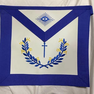 Pode incluir: Avental azul e branco com símbolos bordados. O avental apresenta uma borda azul e um painel central branco com um símbolo de olho azul, uma espada e um desenho de coroa em azul e amarelo.