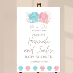 May include: A white baby shower welcome sign on an easel. The sign features blue and pink seashell graphics and the text "He or She WE SHELL SEE!" and "Welcome to Hannah and Joel's BABY SHOWER September 18, 2024."