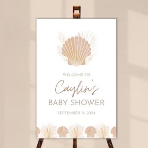 May include: A white welcome sign with a beige seashell and palm leaves. The text reads "Welcome to Caylin's Baby Shower September 18, 2024".