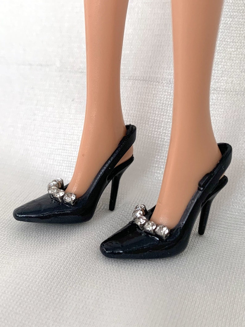 May include: A pair of black heeled shoes with a slingback design. The shoes have a pointed toe and are adorned with a row of clear rhinestones.