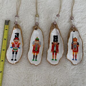 May include: Four white oyster shell ornaments with gold trim, each featuring a different painted nutcracker design. The nutcrackers are wearing red and blue uniforms with gold accents.