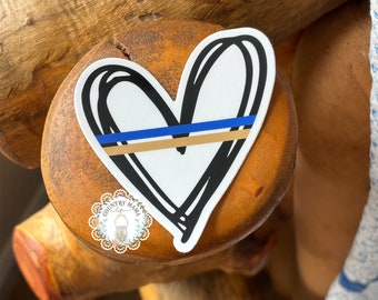 May include: A white heart sticker with a black outline and a thin blue and gold line across the center. The sticker is on a wooden surface.