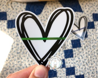 May include: A white heart sticker with a black outline and a green line across the center. The sticker is held in a hand against a blue and white checkered background.