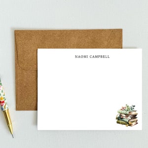 May include: A white stationery card with the name "NAOMI CAMPBELL" printed at the top. The card features a watercolor illustration of a stack of books with green leaves and a few red berries.