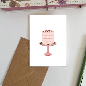 May include: A greeting card featuring a pink cake on a pink cake stand. The cake is decorated with white flowers and cherries. A brown envelope, a pen, and a floral patterned notebook are in the background.