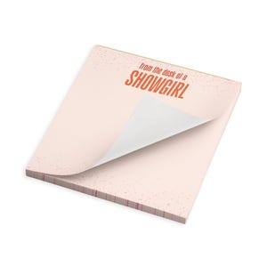 May include: A light pink notepad with the text "from the desk of a SHOWGIRL" in orange. The notepad has a speckled design and a partially lifted top sheet, revealing the white interior pages.