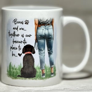 May include: White ceramic mug with a black Labrador Retriever wearing a pink collar, and the text "Bones and me... together is our favourite place to be..." A person in blue jeans and a belt is standing next to the dog.