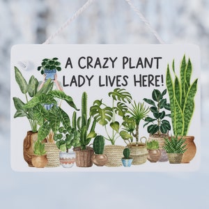 May include: White rectangular sign with the text "A CRAZY PLANT LADY LIVES HERE!" above a watercolour illustration of various potted plants. The sign is suspended by a white string.