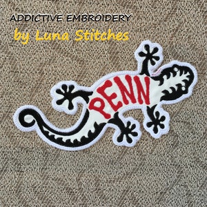 Pode incluir: Etiqueta branca bordada de um gecko preto com a palavra "PENN" em letras vermelhas nas costas. A etiqueta está sobre um fundo de juta marrom.