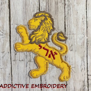 Może przedstawiać: Haftowany lew w kolorze żółtym z brązową grzywą, stojący na tylnych łapach. Lew ma czerwony hebrajski napis na klatce piersiowej.