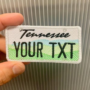 May include: Embroidered patch resembling a Tennessee license plate. The patch features the word "Tennessee" in black script above the text "YOUR TXT" in bold, green letters. A green and blue landscape design is visible.