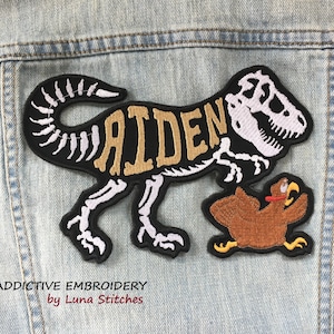 May include: A black and white embroidered patch of a dinosaur skeleton with the name "Aiden" written across its body. The dinosaur is holding a brown embroidered chicken in its mouth. The patch is on a blue denim background. Addictive Embroidery by Luna Stitches is written below the patch.