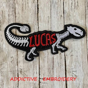May include: Black embroidered patch of a dinosaur skeleton with the name "LUCAS" in red lettering.