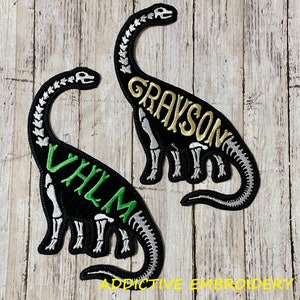 May include: Two black dinosaur skeleton patches with embroidered names. The patch on the left says "VHLM" in green letters. The patch on the right says "GRAYSON" in gold letters. Both patches have silver skeleton details.