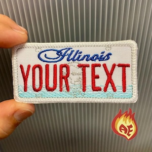 May include: Embroidered patch in the shape of an Illinois license plate. The patch features the word "Illinois" in blue script, the text "YOUR TEXT" in red, and a blue and turquoise water design. A small flame logo is in the bottom right corner.