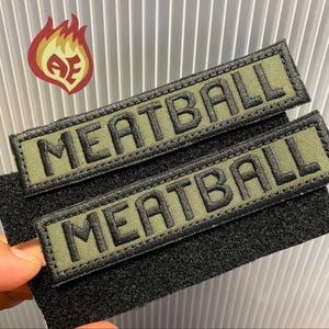 Puede incluir: Dos parches bordados en verde oliva con la palabra "MEATBALL" en hilo negro. Los parches tienen un borde negro y est&aacute;n adheridos a un respaldo de velcro negro. Un logotipo de llama roja, naranja y amarilla se encuentra en la esquina superior izquierda.