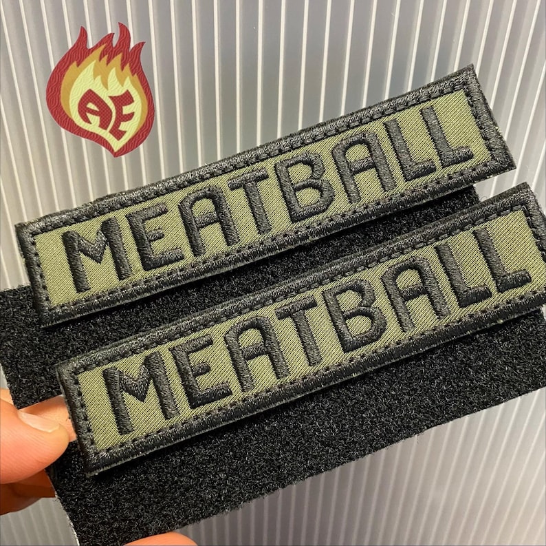 Puede incluir: Dos parches bordados en verde oliva con la palabra "MEATBALL" en hilo negro. Los parches tienen un borde negro y est&aacute;n adheridos a un respaldo de velcro negro. Un logotipo de llama roja, naranja y amarilla se encuentra en la esquina superior izquierda.