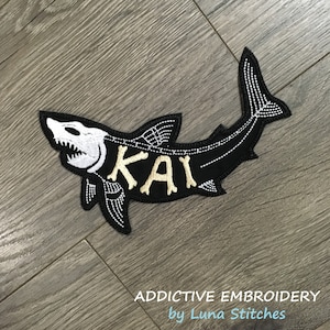 May include: Black embroidered shark patch with white skeleton details and the name "KAI" in gold lettering. The patch is on a woodgrain background. Addictive Embroidery by Luna Stitches.
