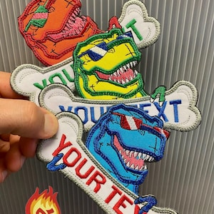 May include: Embroidered patches featuring cartoon dinosaurs wearing sunglasses, each with a bone-shaped banner. The patches are in orange, yellow, and blue, with the text "YOUR TEXT" on the banners. A small patch with flames and the letters "AE" is also visible.