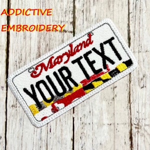 Könnte beinhalten: Ein weißer gestickter Patch mit einem Maryland-Kennzeichen-Design. Der Patch zeigt die Wörter "Maryland" und "YOUR TEXT" in schwarzer Schrift. Der untere Teil des Patches zeigt eine stilisierte Version der Maryland-Staatsflagge in Rot, Gelb und Schwarz.