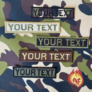 Puede incluir: Parches rectangulares bordados con el texto "YOUR TEXT" en varios colores, incluyendo negro, verde oliva, marr&oacute;n y beige. Los parches se muestran sobre un fondo de camuflaje. Un peque&ntilde;o logotipo de llama es visible.