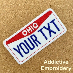 Puede incluir: Parche blanco bordado con borde azul y parte superior roja. El parche tiene forma de matrícula y dice "OHIO" en rojo y "YOUR TXT" en azul.