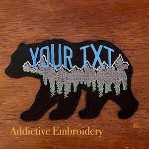 May include: Black embroidered patch of a bear with a mountain range and trees inside the silhouette. The text "YOUR TXT" is embroidered in blue on the bear's back.