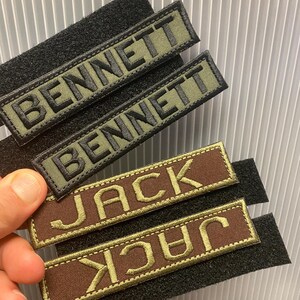 Puede incluir: Parches de nombre bordados con respaldo de velcro. Dos parches dicen "BENNETT" en hilo negro sobre un fondo verde oliva. Dos parches dicen "JACK" en hilo dorado sobre un fondo marr&oacute;n. Los parches son rectangulares.