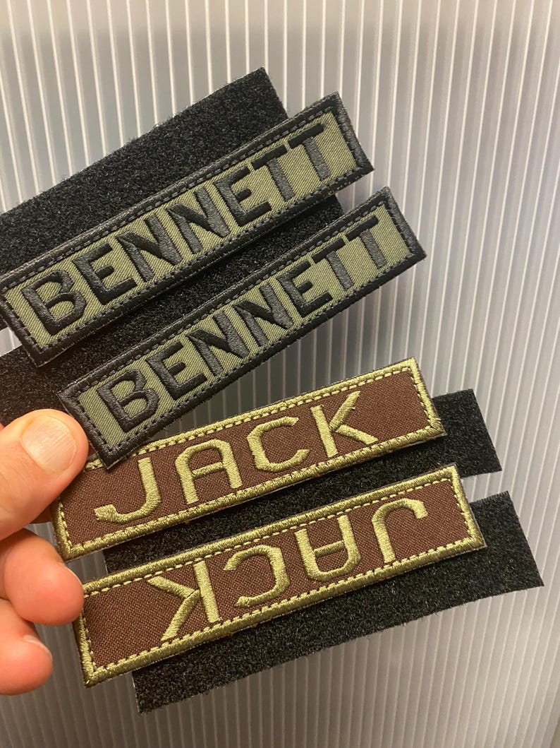 Puede incluir: Parches de nombre bordados con respaldo de velcro. Dos parches dicen "BENNETT" en hilo negro sobre un fondo verde oliva. Dos parches dicen "JACK" en hilo dorado sobre un fondo marr&oacute;n. Los parches son rectangulares.