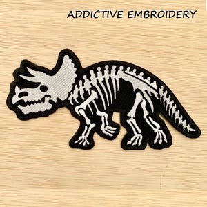 Peut inclure: Patch brodé noir représentant un squelette de tricératops blanc. Le patch porte le texte "ADDICTIVE EMBROIDERY" en haut.