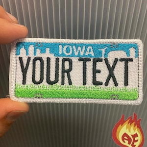 Puede incluir: Parche bordado que se asemeja a una placa de matrícula de Iowa. El parche presenta un cielo azul con una silueta de ciudad, la palabra "IOWA" y el texto "YOUR TEXT" en negro. Hierba verde en la parte inferior. Un pequeño logotipo de llama estilizada en la esquina inferior derecha.