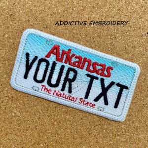 Puede incluir: Parche blanco bordado con un fondo degradado azul y blanco. El parche tiene la forma de una placa de matrícula y dice "Arkansas Your TXT The Natural State" en bordado rojo y negro.