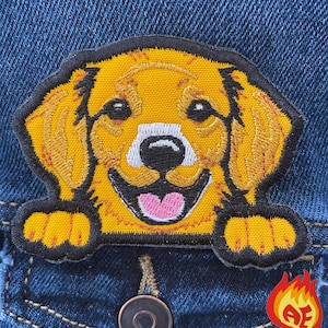 May include: Embroidered patch featuring a smiling golden retriever dog with a black outline. The patch is yellow, with a white muzzle and pink tongue. It is attached to a blue denim jacket.