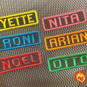 Puede incluir: Seis parches rectangulares bordados con fondo de tela negra y bordes y letras de colores. Los nombres son YETTE en amarillo, NITA en rosa, RONI en azul, ARIAN en naranja, NOEL en rojo y OTTO en verde, todos con un logotipo de llama.