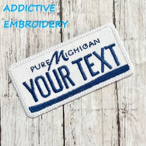 Puede incluir: Parche blanco bordado con costuras azules. El parche tiene forma de matrícula y dice "PURE MICHIGAN" y "YOUR TEXT" en azul.