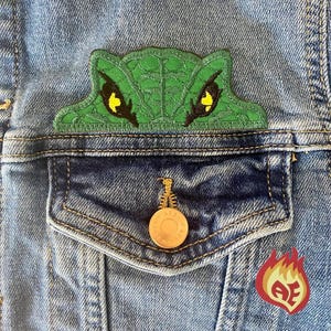 May include: Close-up of a denim jacket with a green embroidered lizard patch above the pocket. The lizard has black and yellow eyes. A gold button and a red and yellow patch are also visible.