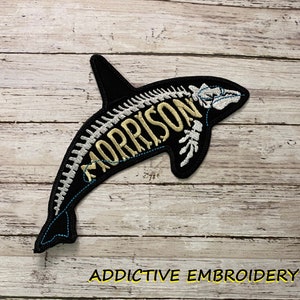 May include: Black embroidered patch of an orca with a skeleton design and the name "MORRISON" written inside the skeleton. The patch is on a wooden background with the text "ADDICTIVE EMBROIDERY" below it.