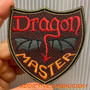 May include: Embroidered shield-shaped patch with the words "Dragon Master" in red and orange thread. The patch features dragon wings and a tail design. The patch is bordered with black and gold thread, with the text "ADDICTIVE EMBROIDERY" at the bottom.