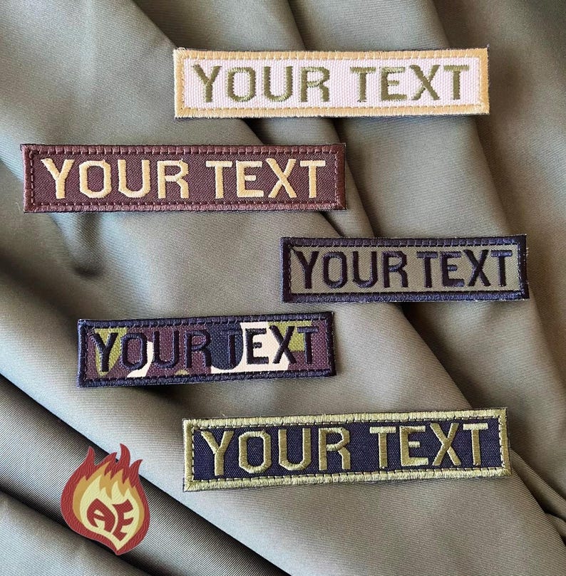 Puede incluir: Parches rectangulares bordados con el texto "YOUR TEXT" en varios colores, incluyendo beige, marr&oacute;n, verde y azul marino. Los parches se muestran sobre un fondo de tela verde. Un peque&ntilde;o logotipo en forma de llama se encuentra en la esquina inferior izquierda.