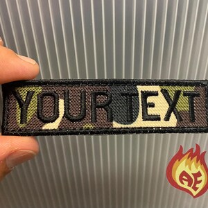 Puede incluir: Un parche rectangular con el texto "YOUR TEXT" bordado en negro sobre un fondo de camuflaje. El parche tiene un borde negro y est&aacute; sujeto por una mano.