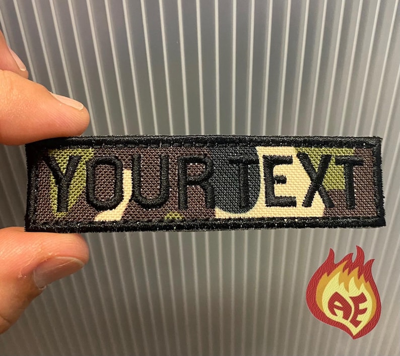 Puede incluir: Un parche rectangular con el texto "YOUR TEXT" bordado en negro sobre un fondo de camuflaje. El parche tiene un borde negro y est&aacute; sujeto por una mano.