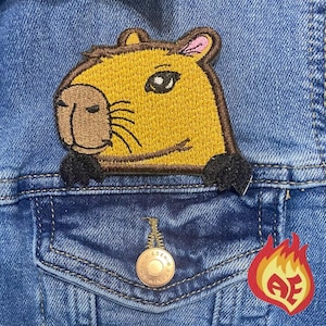 May include: Embroidered patch of a capybara head in shades of yellow, brown, and black, attached to a denim jacket. The patch is detailed with a pink ear and black eyes. A small, red and yellow flame patch is also visible.
