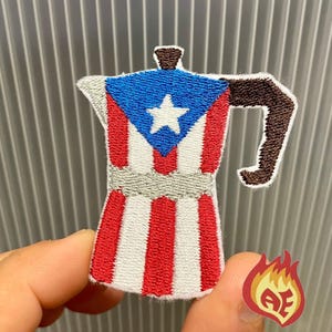Può includere: Toppa ricamata a forma di caffettiera, con la bandiera di Porto Rico. La toppa ha una parte superiore blu con una stella bianca, strisce rosse e bianche e un manico marrone. La toppa è tenuta dalle dita di una persona.