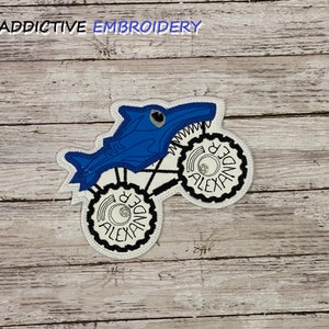 May include: Embroidered patch featuring a blue shark-shaped monster truck design. The truck has white wheels with the name "ALEXANDER" in black lettering. The text "ADDICTIVE EMBROIDERY" is at the top. The patch is on a wooden background.