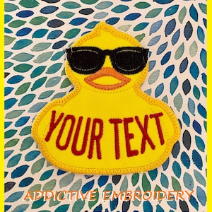 May include: Yellow embroidered patch of a rubber duck wearing sunglasses with the text "YOUR TEXT" in red.